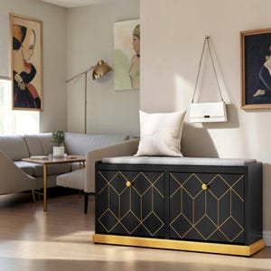 May include: A black rectangular storage bench with two doors featuring a gold geometric line pattern and gold knobs. The bench has a light gray cushioned top and a solid gold base. A white throw pillow rests on the gray seat cushion.