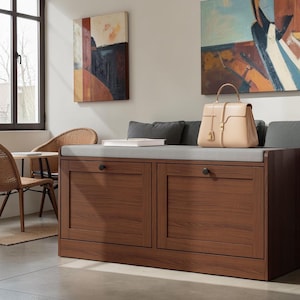 May include: A rectangular wooden storage bench with two front-facing cabinet doors, each featuring a small black circular knob. The bench is topped with a light gray upholstered cushion and sits on a smooth floor, providing a functional furniture piece.