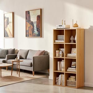 May include: A tall, light-colored wooden bookshelf with eight rectangular cubby compartments. The shelves are filled with various books, small decorative figurines, a white radio, and a woven basket on the bottom right shelf.