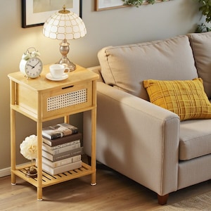 May include: A light wood bedside table with a drawer and a lower shelf. A white alarm clock, a white teacup and saucer, and a lamp with a patterned shade sit on top. A stack of books and a decorative flower are on the lower shelf. A beige sofa and yellow pillow are in the background.