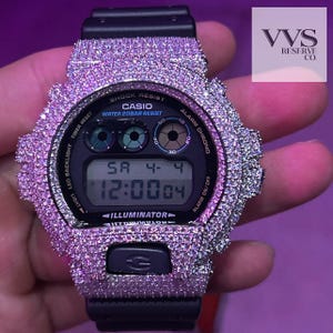 May include: Black Casio digital watch with a bezel and band encrusted with pink gemstones. The watch face displays the time and date. The watch has a black band and a digital display with the text "CASIO" and "ILLUMINATOR".