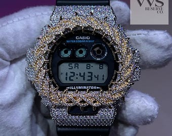 May include: A black Casio digital watch featuring a custom bezel encrusted with numerous clear stones and a gold-toned braided metal accent. The watch face displays the text Casio, Water 20bar Resist, and Illuminator, with a digital time of 12:43:41.