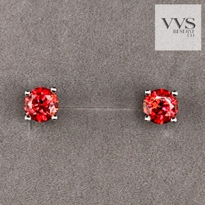 May include: A pair of stud earrings featuring vibrant red gemstones set in a silver metal. The gemstones have a round shape and are mounted on a gray background. The earrings are a classic jewelry item.