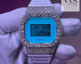 May include: A white Casio digital watch with a light blue face and a sparkling, encrusted bezel. The watch displays the date and time. The band is white. The text "VVS RESERVE CO." is in the upper right corner.