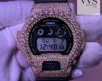 Könnte beinhalten: Eine schwarze Casio Digitaluhr mit einer roségoldenen Lünette und Armband, besetzt mit glitzernden Steinen. Das Zifferblatt zeigt die Uhrzeit, das Datum und den Tag an. Die Uhr hat den Text "CASIO" und "ILLUMINATOR".