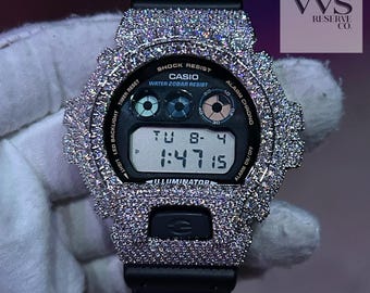 May include: A black Casio G-Shock digital watch with a custom bezel and case fully encrusted in clear, sparkling stones. The watch face displays the text 'SHOCK RESIST', 'CASIO', 'WATER 20BAR RESIST', and 'ILLUMINATOR' with a digital time of 1:47:15.