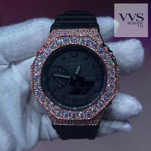 May include: A black G-Shock Casio watch featuring a rose gold-toned octagonal bezel encrusted with numerous clear, sparkling stones. The watch face is black with analog hands and a digital display. The text VVS RESERVE CO. appears in the corner.