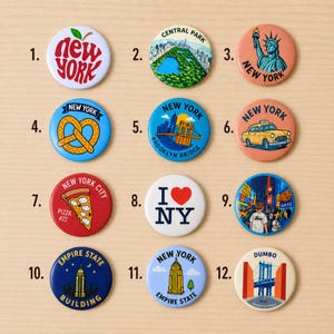 May include: A collection of twelve round New York City themed buttons. Designs include the Statue of Liberty, Brooklyn Bridge, Empire State Building, Central Park, a yellow taxi, and the words "New York" and "I ❤️ NY".