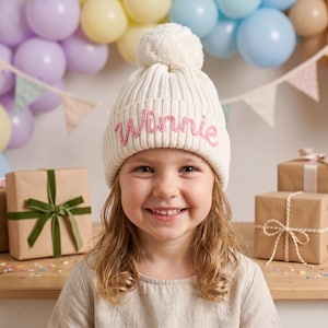 May include: A cream-colored ribbed knit beanie featuring a large pom-pom on top and the name Winnie embroidered in pink cursive script on the folded cuff. The hat is made of a thick, textured yarn and has a classic winter style design.