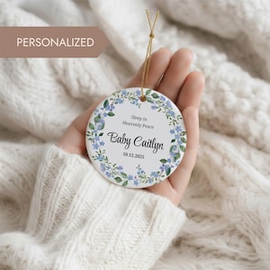 Pode incluir: Enfeite de cerâmica personalizado com design floral. O enfeite apresenta o texto "Sleep in Heavenly Peace" e "Baby Caitlyn 10.12.2025". É branco com detalhes florais azuis e verdes, e um cordão dourado para pendurar.