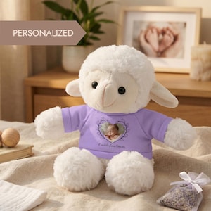 Könnte beinhalten: Ein weißes Plüschlamm mit einem lila Shirt mit einem Babyfoto in einem herzförmigen Design. Auf dem Shirt steht "A cuddle from Heaven". Das Lamm sitzt auf einer beigen Decke. Das Wort "PERSONALISIERT" steht in einem Banner oben links.