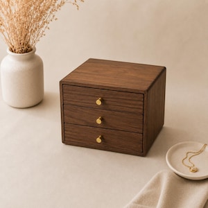 Könnte beinhalten: Eine Aufbewahrungskommode aus dunkelbraunem Holz mit drei horizontalen Schubladen, die jeweils mit einem kleinen, runden, goldfarbenen Metallknopf versehen sind. Die Kommode hat einen glatten, rechteckigen Rahmen und steht auf einer ebenen Fläche.