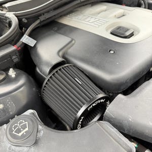 Puede incluir: Un filtro de aire cilíndrico negro con material plisado y una tapa metálica negra con el texto blanco PRORAM instalado en el compartimento del motor de un vehículo. El filtro está colocado junto a una cubierta de motor de plástico negro.