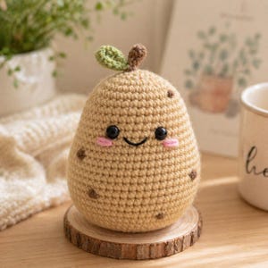 May include: A crocheted potato toy with a smiling face, black eyes, and pink cheeks. The tan-colored potato has brown spots and a green leaf and brown stem on top. It sits on a small wooden slice.