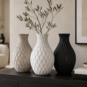 May include: Three decorative vases in neutral tones. The vases are arranged on a dark surface, with a light beige, white, and black vase. Each vase has a textured diamond pattern. A sprig of greenery is in the white vase.