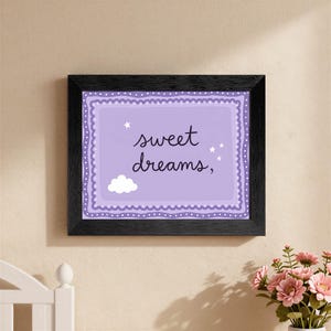 May include: A framed print with the words "sweet dreams," in cursive on a lavender background. The print has a white cloud and stars, and is framed in a black frame. The print is hanging on a beige wall.