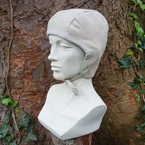 Könnte beinhalten: Eine hellbeige gesteppte Mütze mit weichem Rand und Kinnbändern, präsentiert auf einer weißen Büste. Die Mütze hat ein strukturiertes Aussehen und eine dezente, neutrale Farbpalette. Der Hintergrund zeigt einen Baumstamm und grünes Laub.