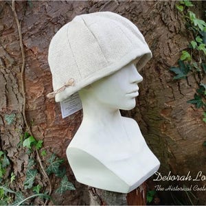 Puede incluir: Un sombrero de lino beige con una corona redondeada y un borde que se curva hacia abajo alrededor de las orejas. El sombrero está hecho de un tejido tejido y tiene un diseño simple.