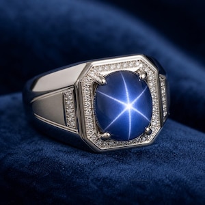 Può includere: Un anello d'argento con una grande gemma blu ovale che mostra un motivo a stella a sei raggi. La gemma è circondata da un alone di piccole pietre trasparenti. L'anello ha un design rettangolare con piccole pietre sui lati.