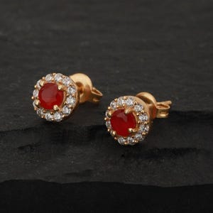 May include: A pair of gold-colored stud earrings. Each earring features a round, red gemstone surrounded by a halo of small, clear gemstones. The earrings are set against a dark, textured background.