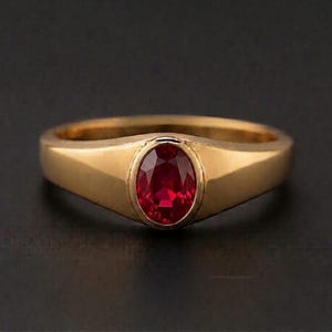 May include: A gold ring featuring a large, oval-cut ruby gemstone. The ring has a simple, classic design with a polished finish. The ruby is a deep red colour and is the focal point of the ring.