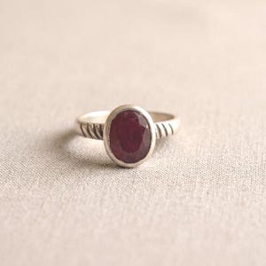 May include: A silver ring with an oval, deep red gemstone. The band has a textured design on each side of the gem. The ring is set against a neutral, textured background.