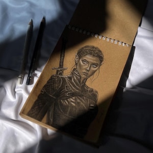 May include: A pencil sketch of a woman with pointed ears, wearing armor, and holding a sword, in a spiral-bound sketchbook. Two pencils are visible to the left of the sketchbook. The drawing is in shades of gray and brown.