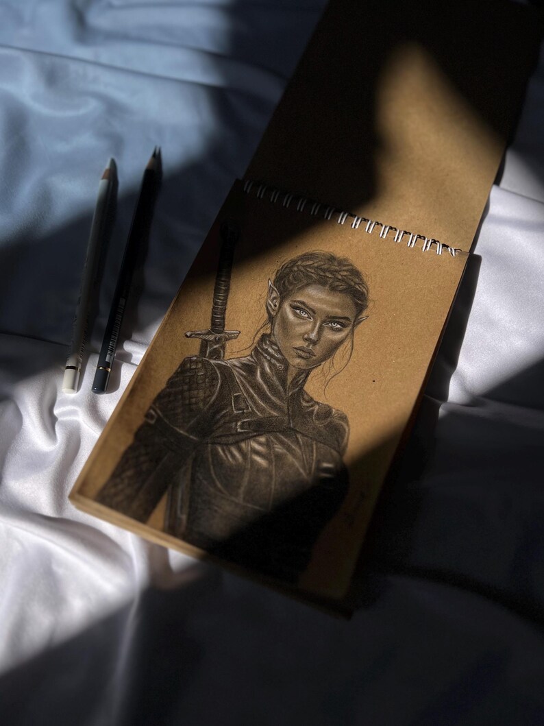 May include: A pencil sketch of a woman with pointed ears, wearing armor, and holding a sword, in a spiral-bound sketchbook. Two pencils are visible to the left of the sketchbook. The drawing is in shades of gray and brown.