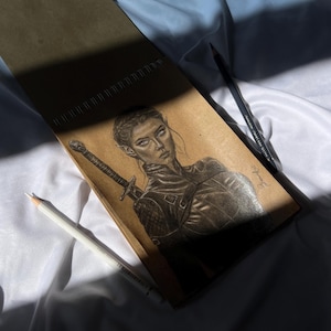 May include: A pencil sketch of a woman with pointed ears, holding a sword, in a leather-like outfit. The drawing is on a brown spiral-bound sketchbook, with a white and a black pencil resting on a white fabric.