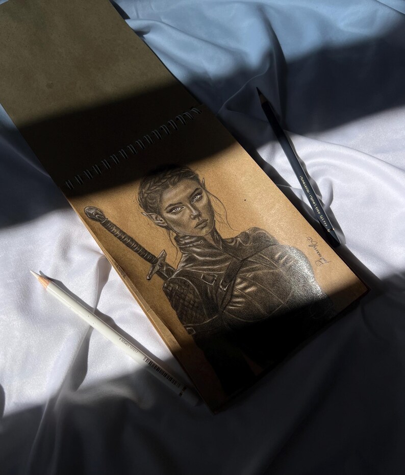 May include: A pencil sketch of a woman with pointed ears, holding a sword, in a leather-like outfit. The drawing is on a brown spiral-bound sketchbook, with a white and a black pencil resting on a white fabric.