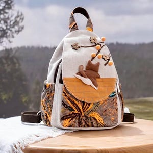 May include: A beige canvas backpack with a brown and orange floral pattern. The backpack features a brown cat applique on the front flap, along with decorative leaves and orange pom-poms. The bag has a brown handle and straps.