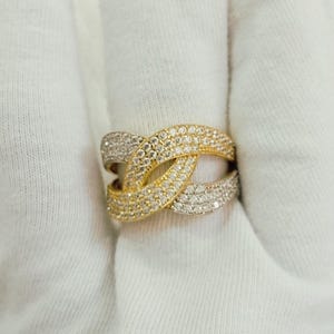 May include: A wide band ring featuring an intricate crossover design. The ring is crafted with alternating sections of yellow metal and silver-toned metal, both fully encrusted with numerous small, clear, sparkling stones.