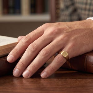 May include: A gold signet ring with a smooth, matte finish and a flat, square-shaped face. The center of the ring face features a single, engraved letter C in a cursive script style. The band is solid gold and maintains a consistent width.
