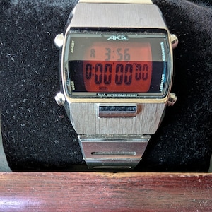 May include: A silver-toned digital wristwatch with a brushed metal finish and a rectangular case. The watch face features a red digital display showing the time 3:56 and a stopwatch function. The text AKA and ALBA WATER 10BAR RESIST are visible.