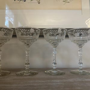 Può includere: Un set di quattro coppe in vetro trasparente con un delicato motivo floreale e a foglie inciso sul bordo. Ogni bicchiere presenta una coppa ampia e poco profonda e uno stelo decorativo sfaccettato che poggia su una base circolare.