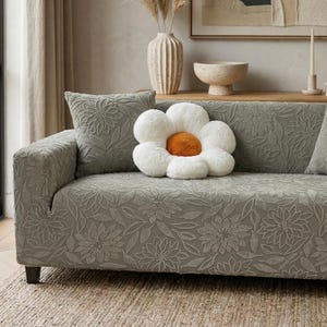 May include: A gray sofa with a floral pattern, a matching pillow, and a large flower-shaped pillow with a white and orange design. The sofa sits on a woven rug, with a vase and artwork in the background.