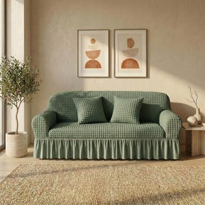 May include: A sage green sofa with two matching square cushions. The sofa has a textured pattern and a ruffled skirt. Two framed abstract art prints hang above the sofa, and a small olive tree is to the left.