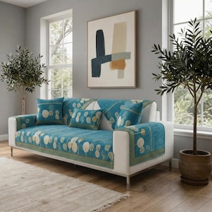 Op de afbeelding: Een witte bank met een turquoise patroonhoes en bijpassende kussens. De hoes heeft een boomontwerp met gouden accenten. De bank staat in een kamer met een neutraal kleurenpalet, een groot schilderij en twee potbomen.