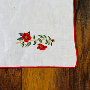 May include: White linen placemat with a red embroidered floral design. The placemat has a red border.