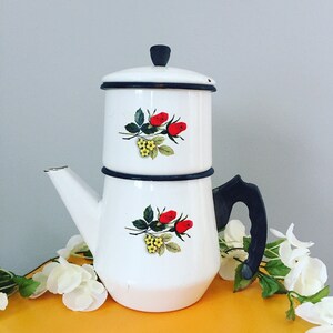 May include: White enamel coffee pot with a black handle and a floral design of red roses and green leaves. The pot has a separate top section for brewing coffee.