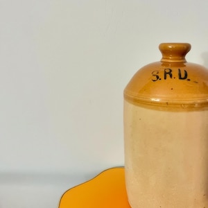 Może przedstawiać: Antyczny ceramiczny dzban z brązową glazurą i białą podstawą. Dzban ma szeroki otwór i zaokrąglone ciało. Dzban jest oznaczony "S.R.U." czarnymi literami.