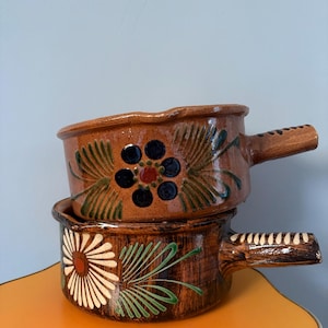 Puede incluir: Dos cuencos de cerámica marrón apilados con asas. El cuenco superior presenta un diseño floral con detalles azules, rojos y verdes. El cuenco inferior tiene un diseño de margarita blanca y roja con hojas verdes.