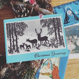 Può includere: Un biglietto di Natale blu con la silhouette di una famiglia di cervi in un ambiente boschivo. Il biglietto recita "Christmas Greetings".