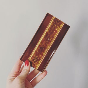 May include: A brown and yellow ceramic tile with a textured surface. The tile has a brown border and a yellow center with a speckled pattern.