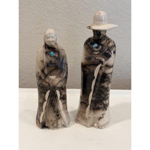 May include: Two standing figurines crafted from smooth, off-white material with black horsehair-style markings. Each figure features a small, round, bright blue turquoise stone inlay at the neckline. One figure wears a wide-brimmed hat.