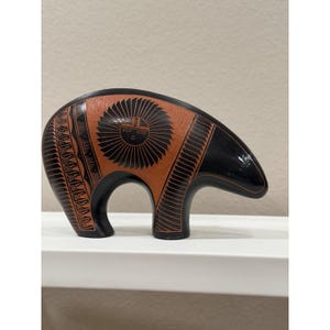 May include: A ceramic bear figurine featuring a matte terracotta-colored body with black painted details. The bear displays a sun-like circular motif on its side, surrounded by intricate black geometric patterns and solid black head and leg sections.