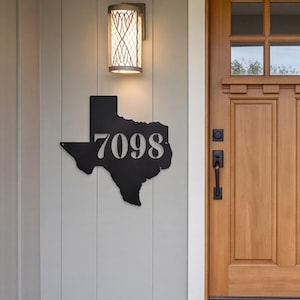 May include: A matte black metal wall sign shaped like the state of Texas featuring the house numbers 7098 cut out in a bold, sans-serif font. The sign is mounted on a light gray vertical paneled wall next to a wooden door and a wall light.
