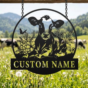 May include: Black metal cow sign with a circular design, featuring a cow resting among flowers and a butterfly. The sign hangs from chains and includes a rectangular area with the text "CUSTOM NAME". The background shows a green field and mountains.