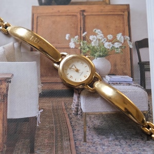 Könnte beinhalten: Eine goldfarbene Armbanduhr mit rundem Zifferblatt und Gliederarmband. Das Zifferblatt hat einen weißen Hintergrund mit schwarzen Ziffern und Zeigern. Das Armband und das Uhrengehäuse sind goldfarben glänzend. Die Uhr liegt auf einer Zeitschrift.
