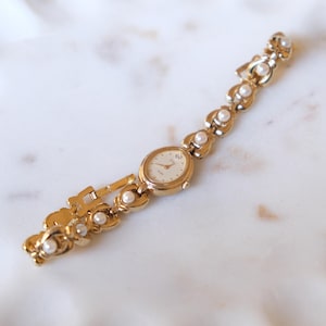 May include: A gold-tone wristwatch featuring an oval-shaped face with a light-colored dial and gold hands. The bracelet is composed of interconnected gold-tone links, each accented with a small, round white pearl, creating a continuous decorative pattern.